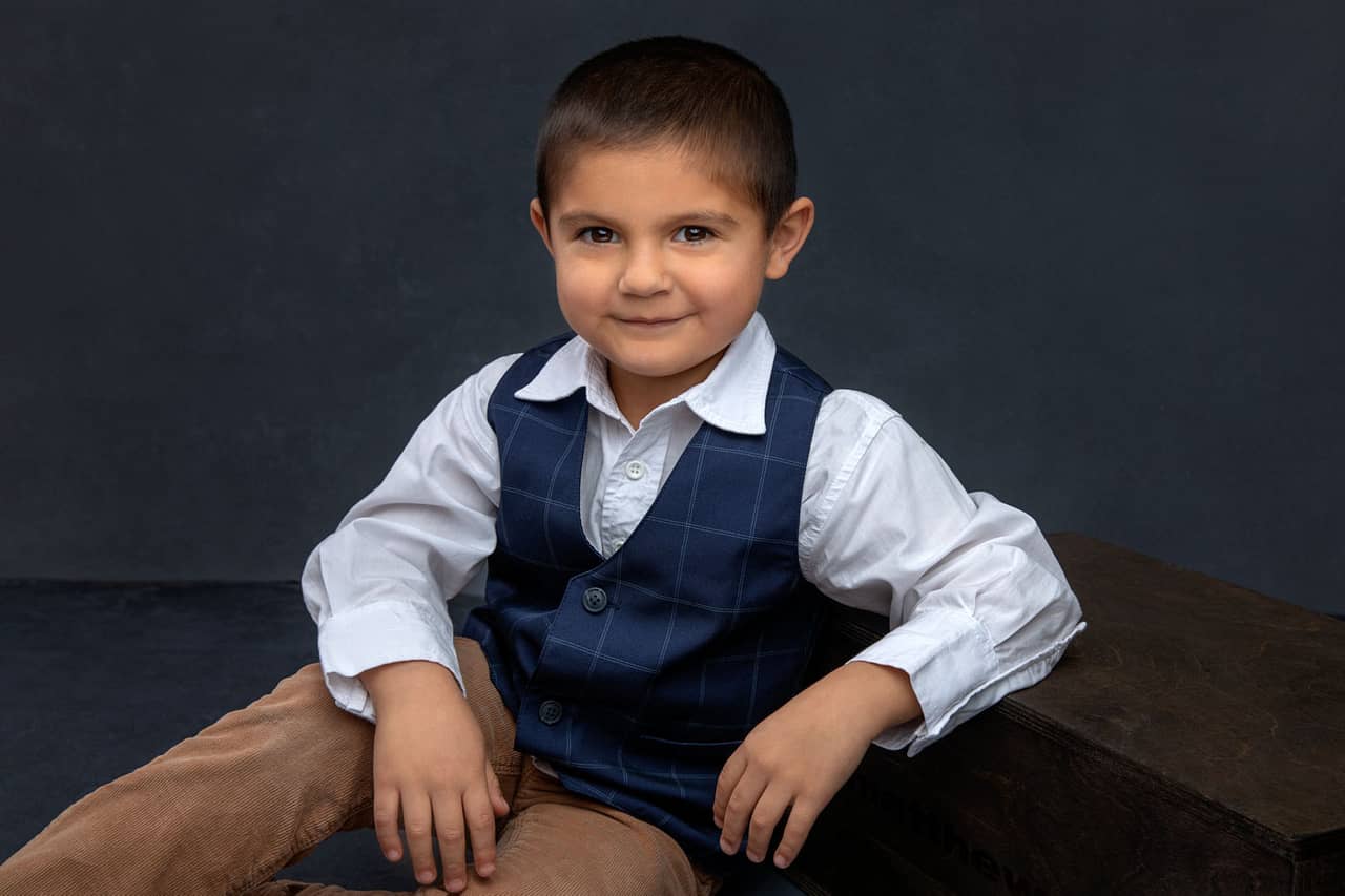 Cute photo of an adorable little boy smirking and sitting with his elbow resting on a box. He's wearing a white dress shirt with a blue plaid vest and tan pants. Josh and Stefanie Fraley Family Portrait Photography.