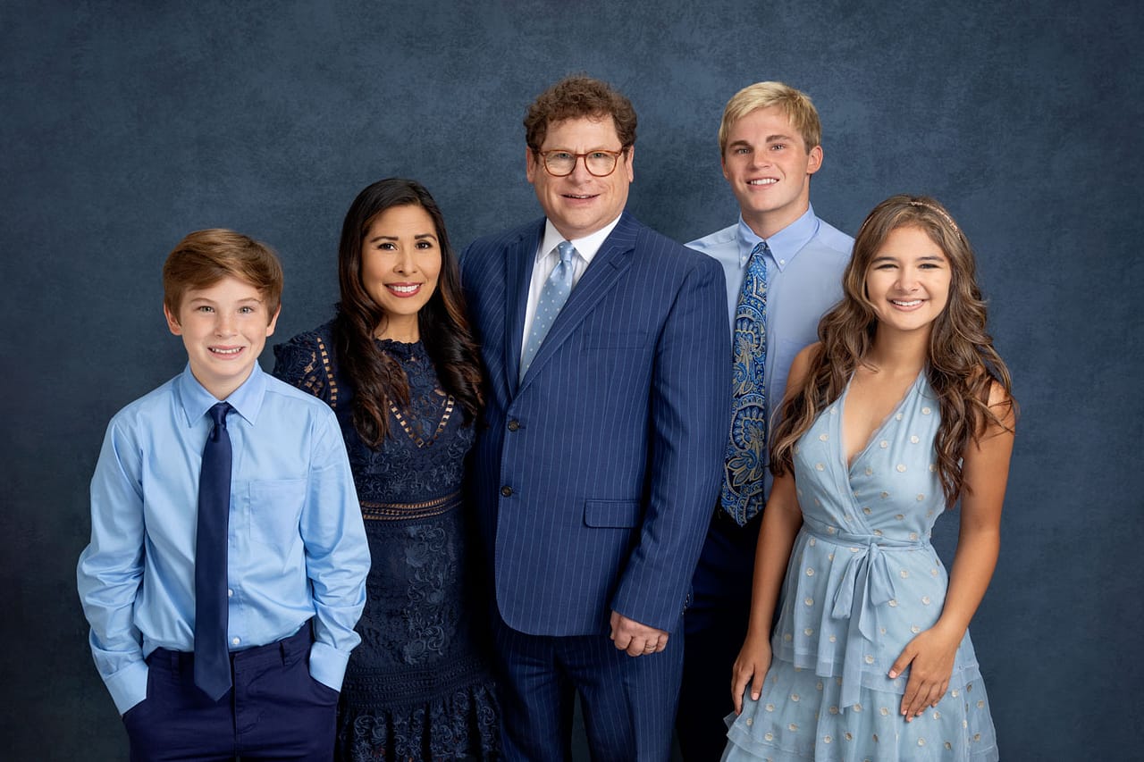 Precious photo of a cute, smiling family standing close. All are wearing shades of blue on a blue backdrop. Guy Furay and Myra Ruiz Family Portrait Photography.