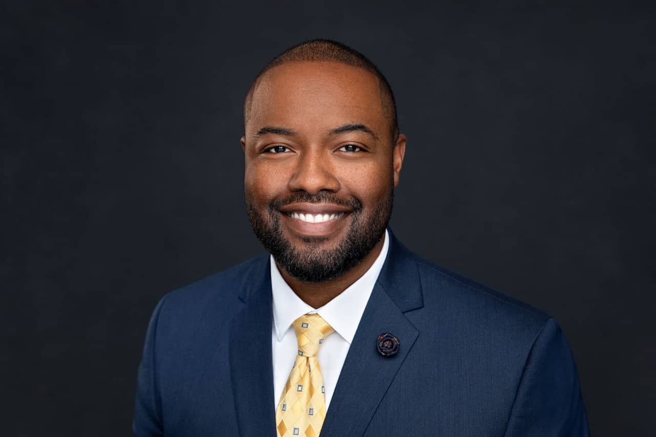 Professional headshot photo of a handsome, smiling black man wearing a white shirt, a yellow tie and a blue jacket with a rose lapel pin. Shannon Windham Headshot Photography.