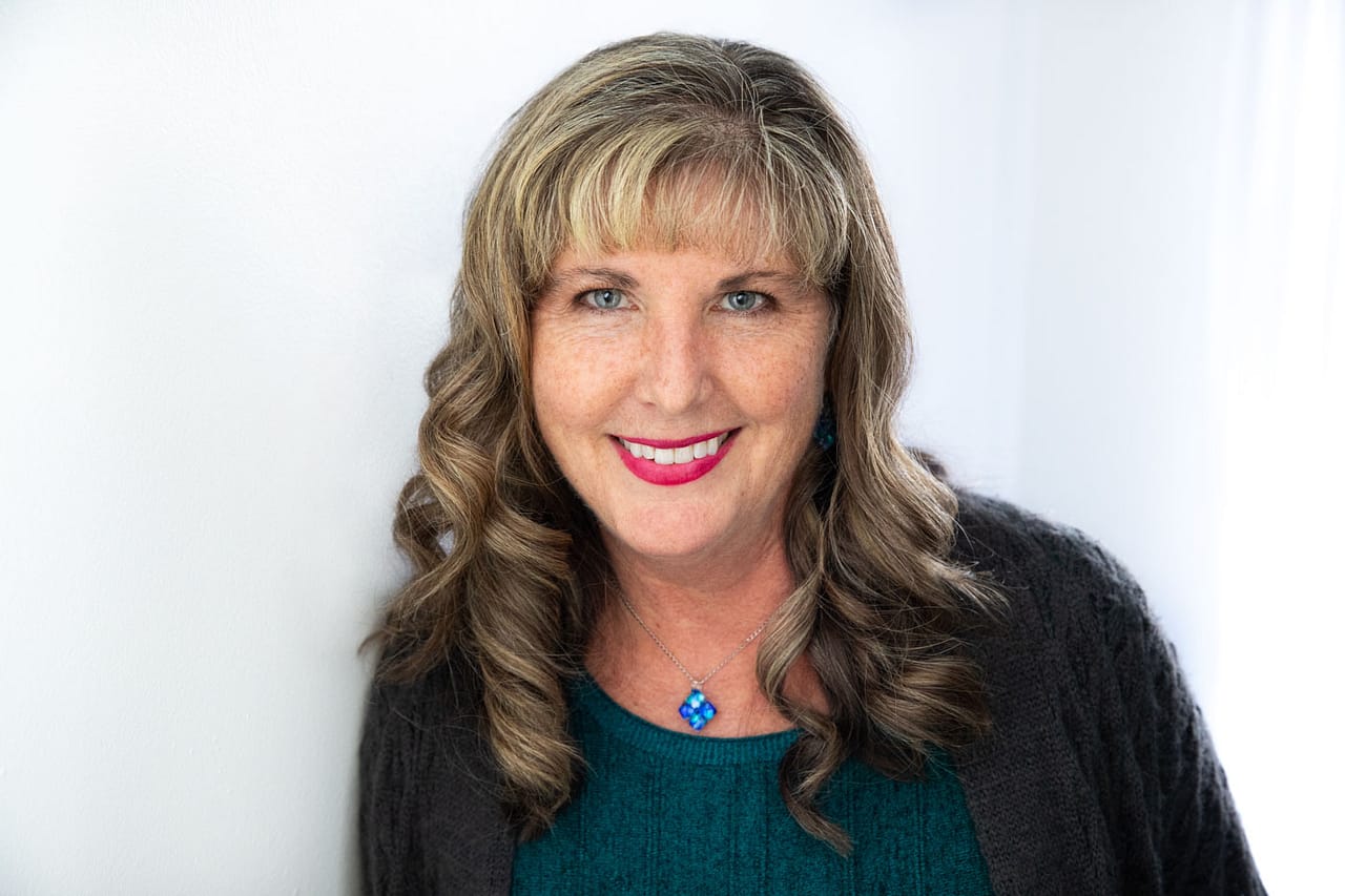 Professional headshot photo of a pretty, smiling woman with dirty blonde hair and red lipstick. She's wearing a green shirt with a gray sweater and a necklace with a blue penant. Shirley Sepulveda Headshot Photography.