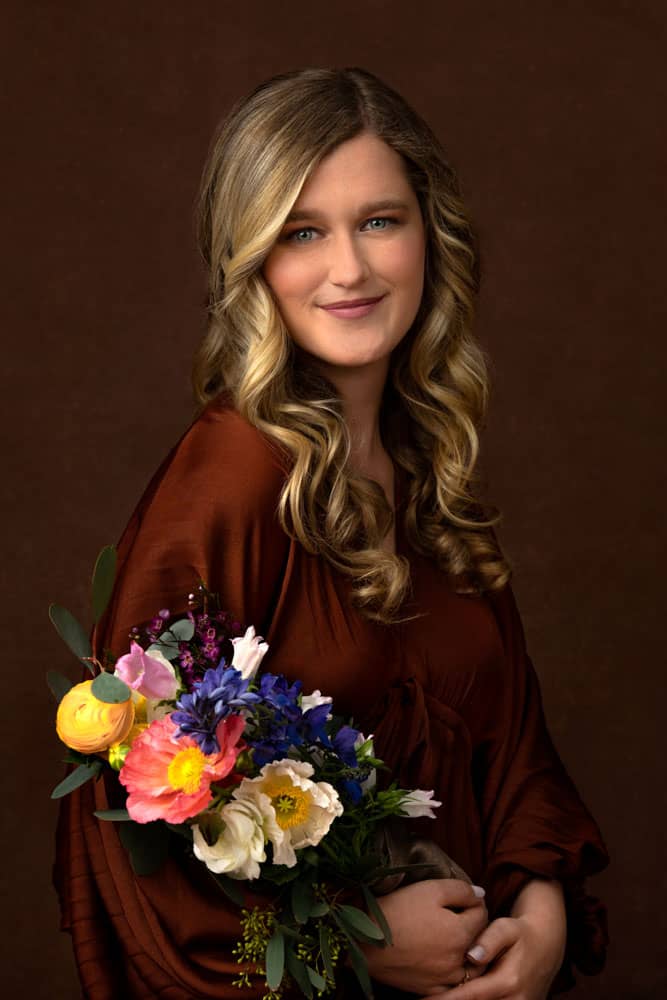 Moody image of a pretty, smiling young woman with blond hair and brown dress on brown backdrop. She's holding pink, yellow, white and blue flowers. Portrait photography session with Printed Water and Alicia Waller.