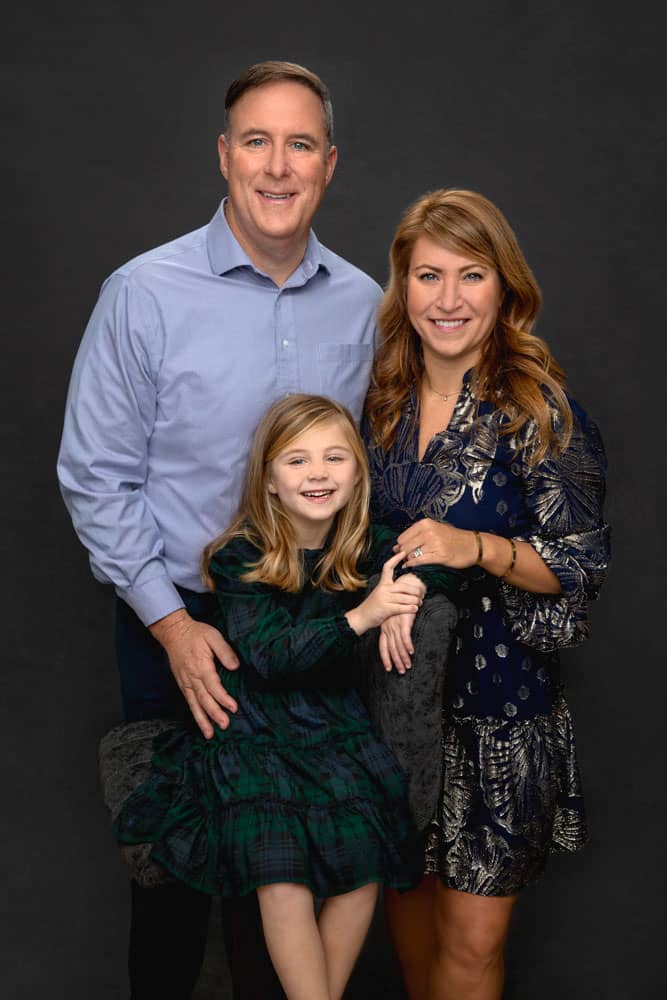 Precious photo of beautiful family of three cuddled close together. Father, mother and daughter are all whearing blues and greens. They are set on a dark backdrop. Chris and Amber Edwards Family Portrait Photography.