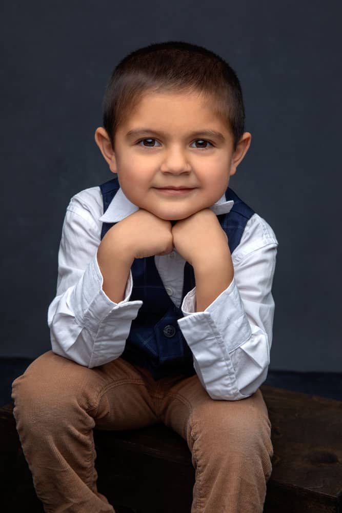 Cute photo of an adorable little boy sitting on a box with his elbows resting on his knees and his chin resting on his hands. He's wearing a white dress shirt with a blue plaid vest and tan pants. Josh and Stefanie Fraley Family Portrait Photography.