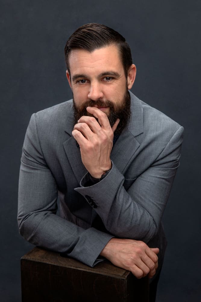 Sharp photo of a handsome bearded man with a gray jacket leaning on box, his hand touching his beard and mouth. Josh and Stefanie Fraley Family Portrait Photography.