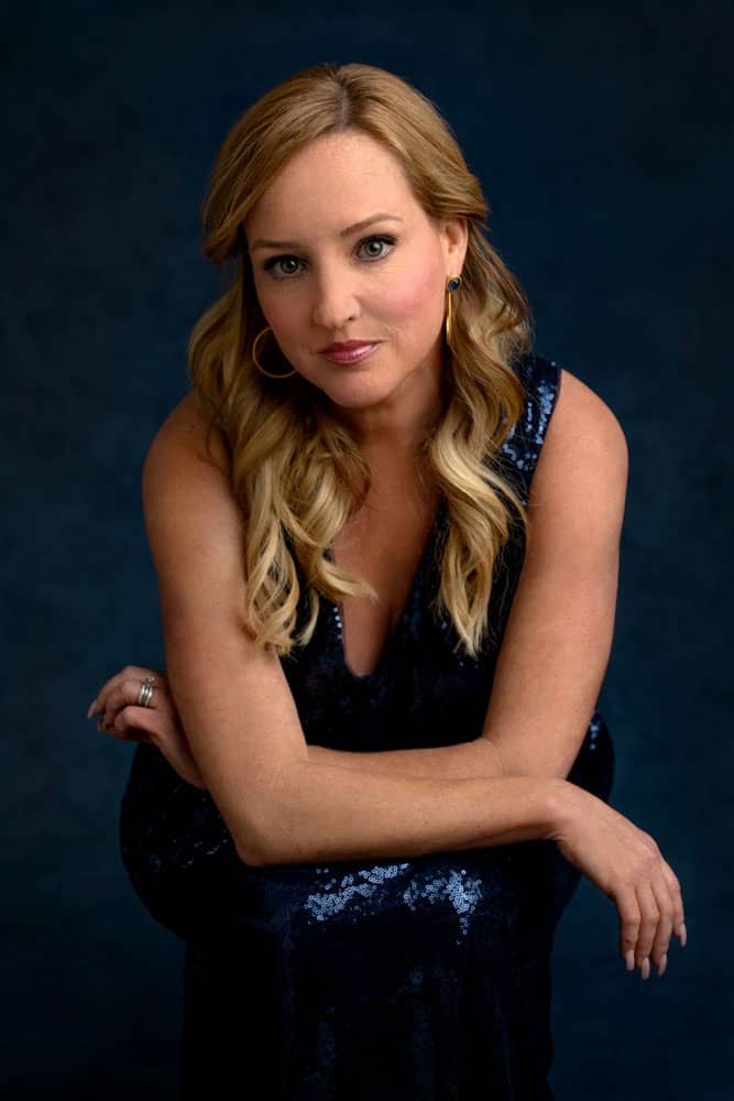 Moody, classy photo of a pretty young woman sitting, here arms resting on her knees. She's wearing a dark blue siquen dress and set on a dark blue backdrop. Meredith Dowless Women's Portrait.