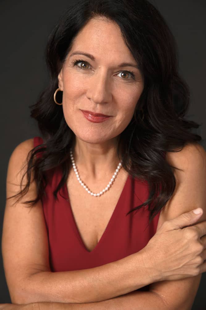 Classy professional photo of a woman with long dark brown hair. She is wearing a red shirt, a pearl necklace, earrings and red lipstick. She is holding her arm with one hand and leaning on the other.
