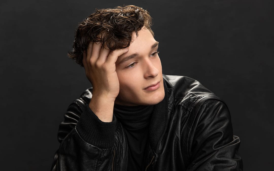 Sharp photo of a handsome young man with curly brown hair looking away wearing a black leather jacket. His head is leaning on his hand, with his fingers in his hair. Lucas Nichter Senior Portrait Photography.
