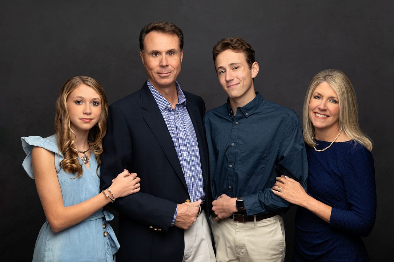 Cute photo of a happy family of four smiling and standing close. All are wearing shades of blue and standing on a dark backdrop. Don and Beverly Elmore Family Portrait Photography.