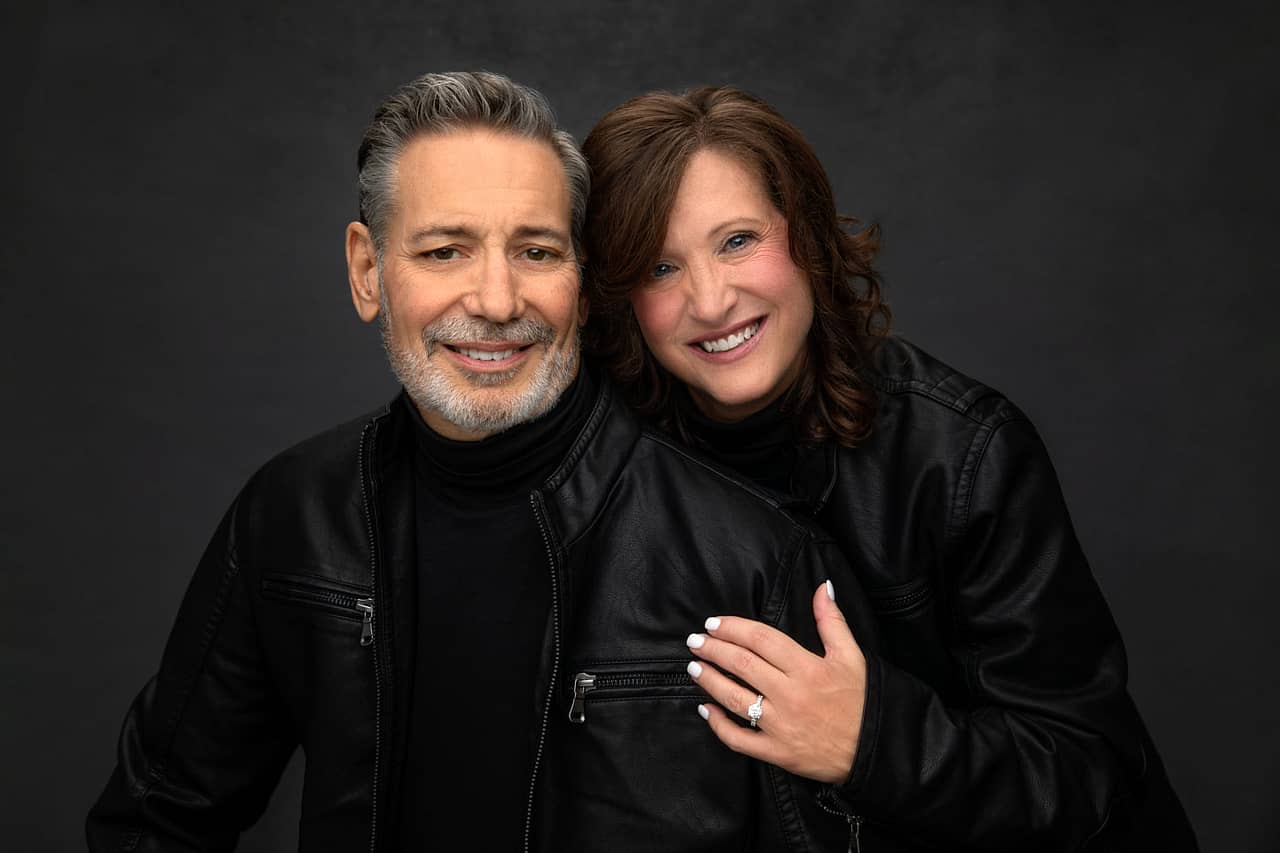 Cute photo of handsome man with attractive woman, both wearing black jackets. She's holding his arm, showing her new engagement ring. Donald and Charlotte Aucoin engagement portrait photography.