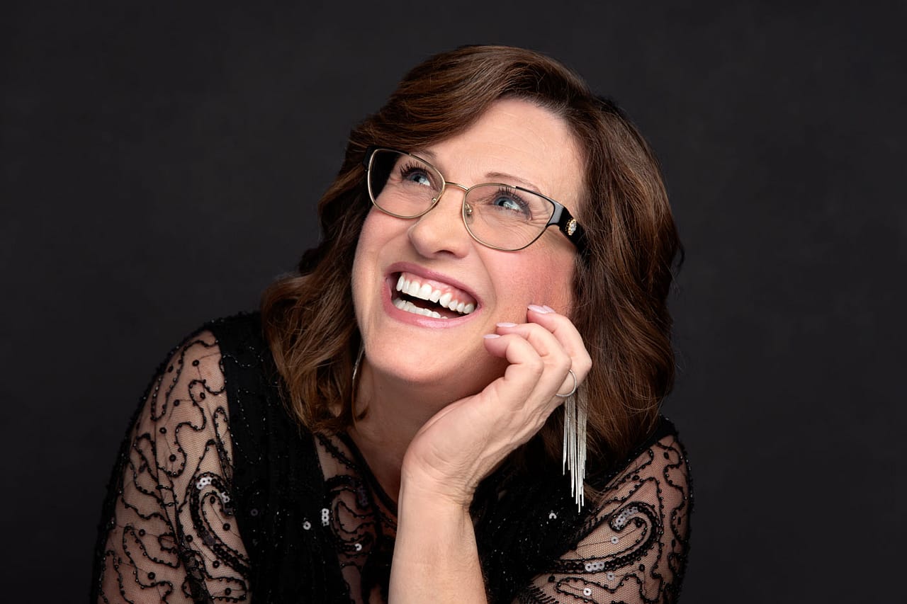 Joyful photo of a pretty woman looking up and smiling. She's wearing a black top, long earrings and glasses. She's set on dark backdrop. Charlotte Aucoin Personal Branding Photography.