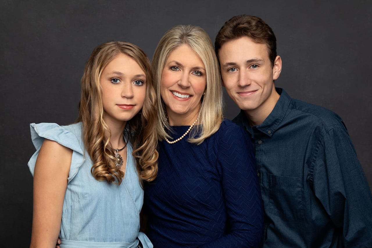 Cute photo of a mother, son and daughter standing close and smiling. They are all wearing blue on a dark backdrop. Don and Beverly Elmore Family Portrait Photography.