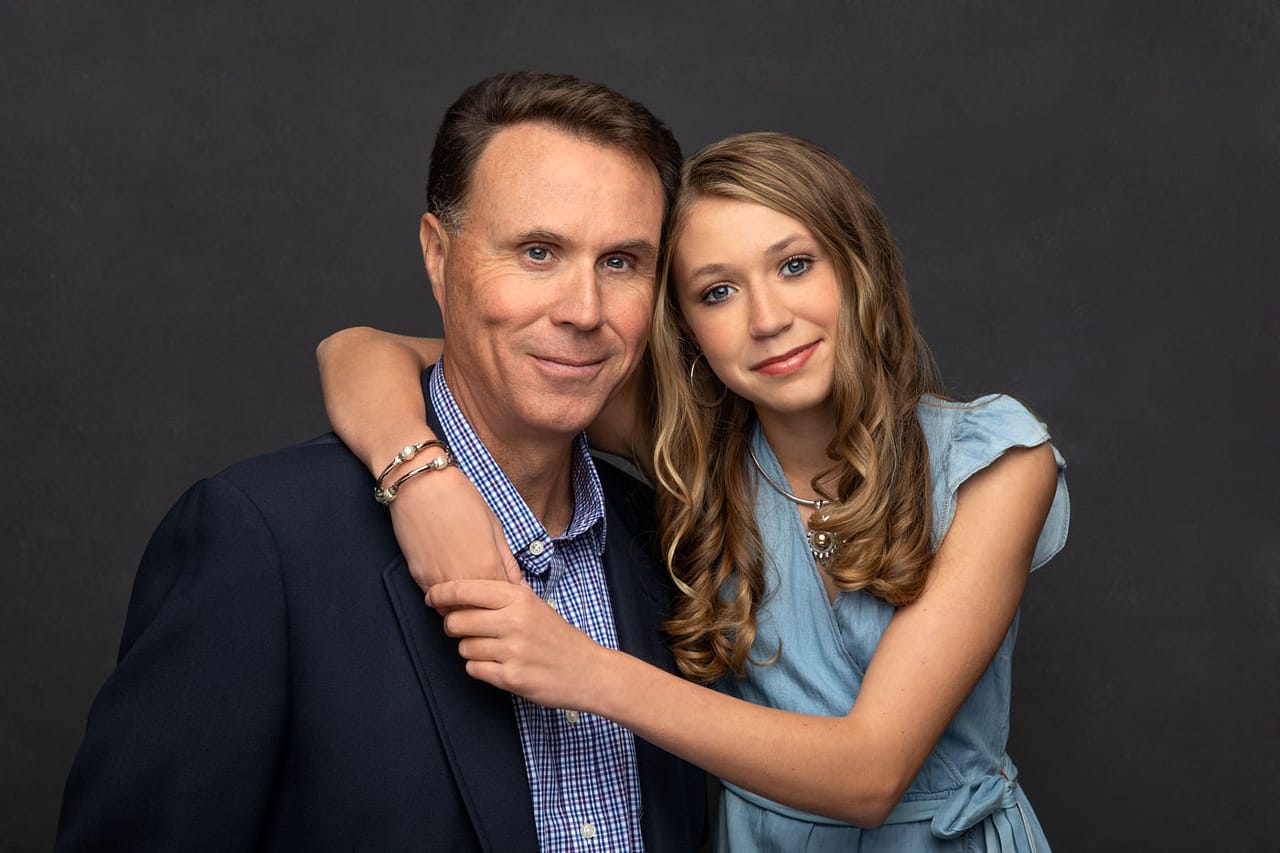 Precious photo of a father and daughter standing close, her arm around his shoulder. Both wearing blue on a dark backdrop. Don and Beverly Elmore Family Portrait Photography.