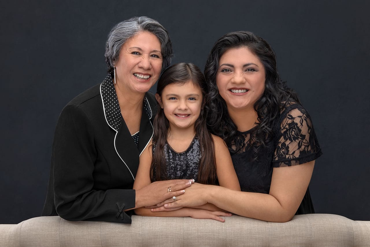 Beautiful generational portrait of smiling grandmother, mother and daughter. They are standing, leaning on the back of a couch with their hands placed on top of each other hand. All three are wearing black. Josh and Stefanie Fraley Family Portrait Photography.
