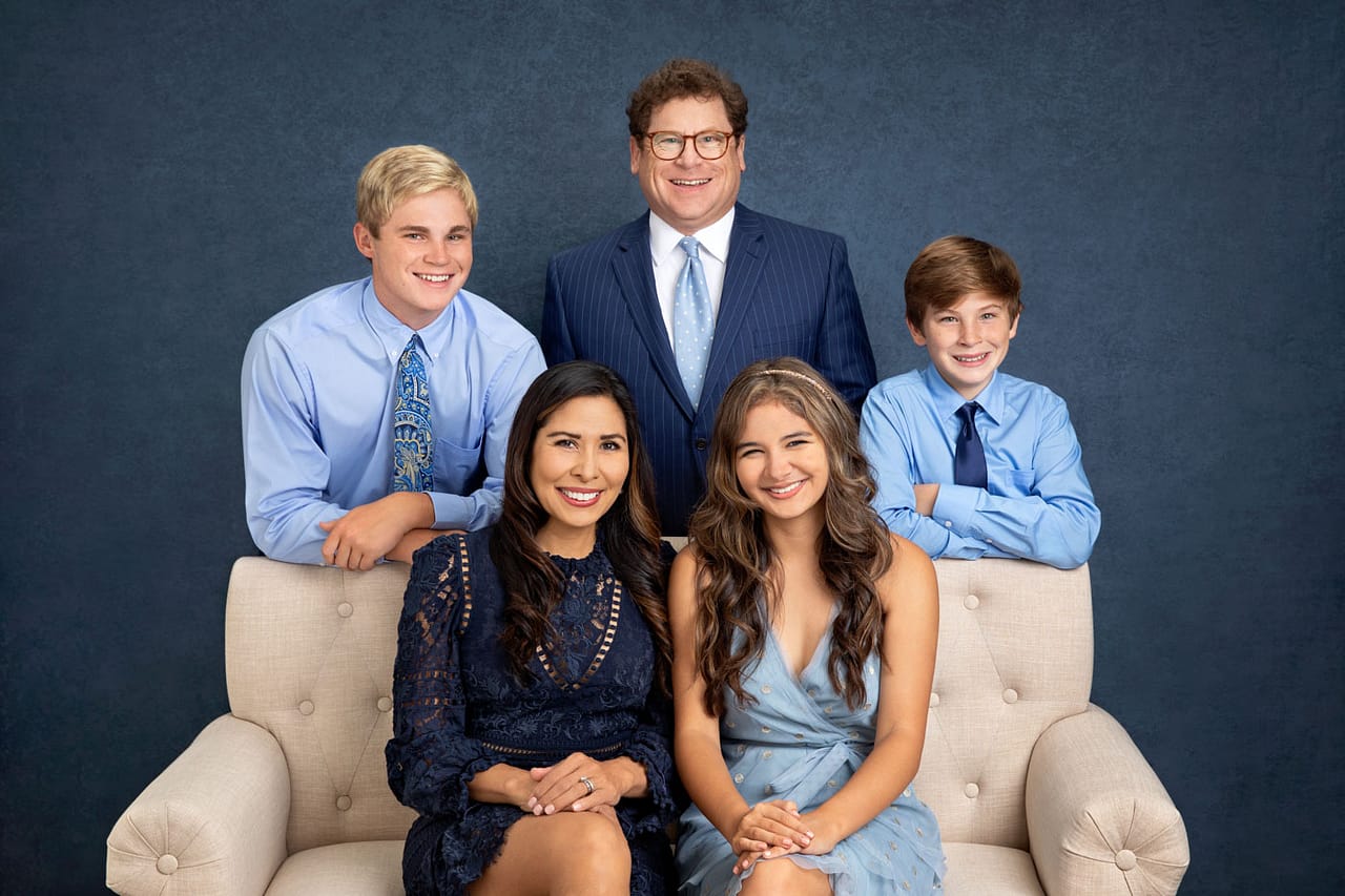 Precious photo of a cute, smiling family sitting on and standing around a couch. The men are leaning on the couch, and the women are sitting. All are wearing shades of blue on a blue backdrop. Guy Furay and Myra Ruiz Family Portrait Photography.