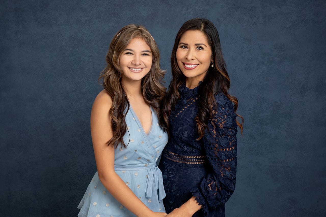 Precious photo of a smiling mother and daugther standing close holding hands. Both are wearing blue dresses. Guy Furay and Myra Ruiz Family Portrait Photography.