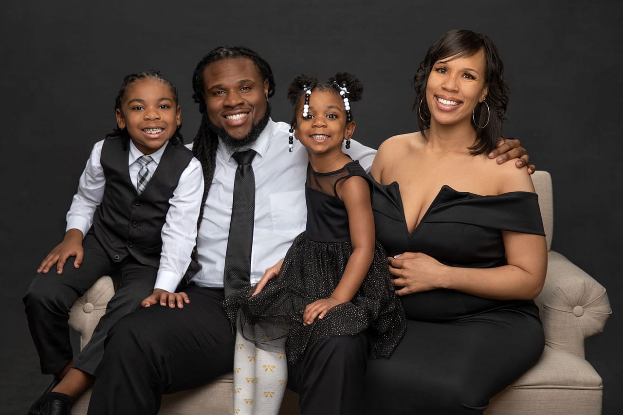 Beautiful maternity and family photo of precious family sitting close to each other on a tan couch. The family is dressed in black and white, with the little boy wearing a black vest with his tie. Dad is wearing a black tie and mom and the daugther are wearing a black dress. Holmes Family Portrait Photography.