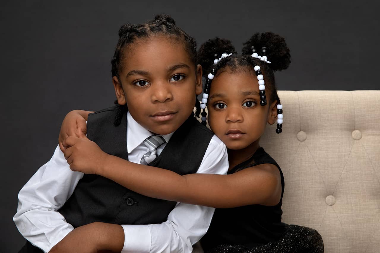 Precious photo of cute brother and sister. Brother is leaning on a tan couch, while sister is sitting on the couch, with her arms around him, holding him close. She is wearing a black dress and he is wearing a white shirt with a black vest. Holmes Family Portrait Photography.