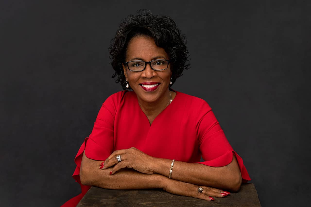 Classy photo of a pretty, smiling black woman with glasses wearing a bright red shirt, black necklace and golden earrings. She's wearing red lipstick and red nails. Janice Butler Women's Portrait Photography.