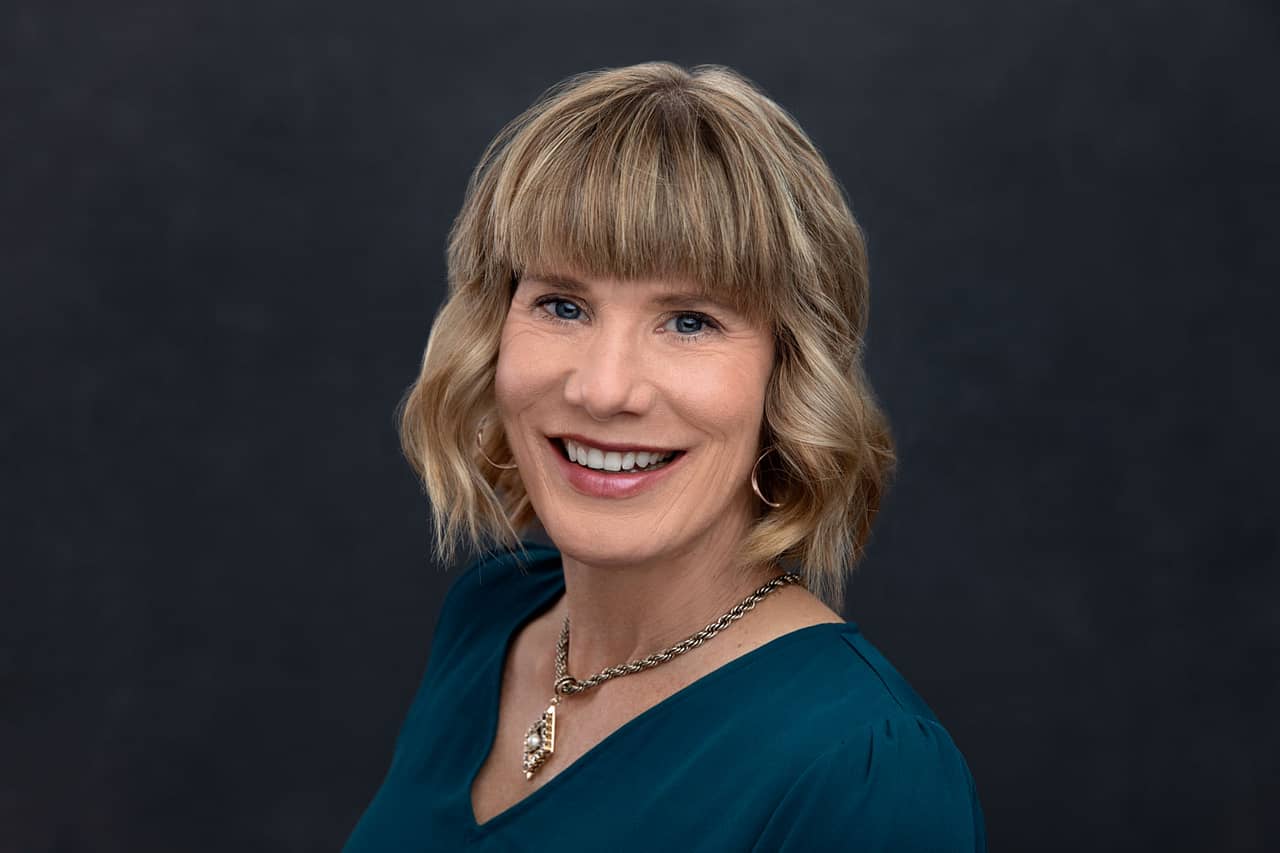 Professional headshot of a pretty, smiling woman with short dirty blonde hair. She is wearing red lipstick, a blue shirt and a necklace. Jody Whitaker Headshot Photography.