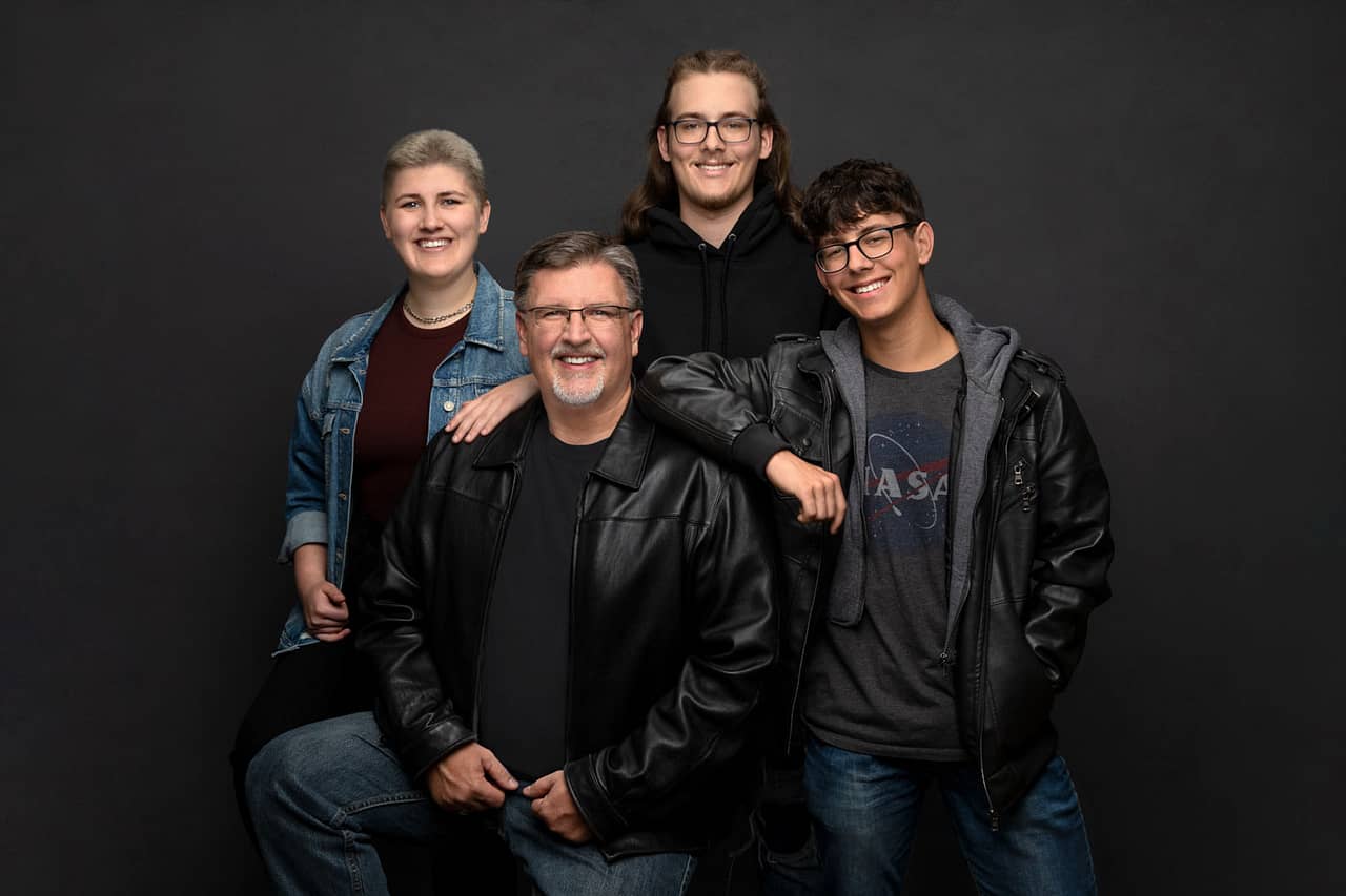 Family photo of smiling, happy family wearing black and blue. A father and three teenage children, two of them leaning on him as he sits down. James Anderson Family Portrait Photography.