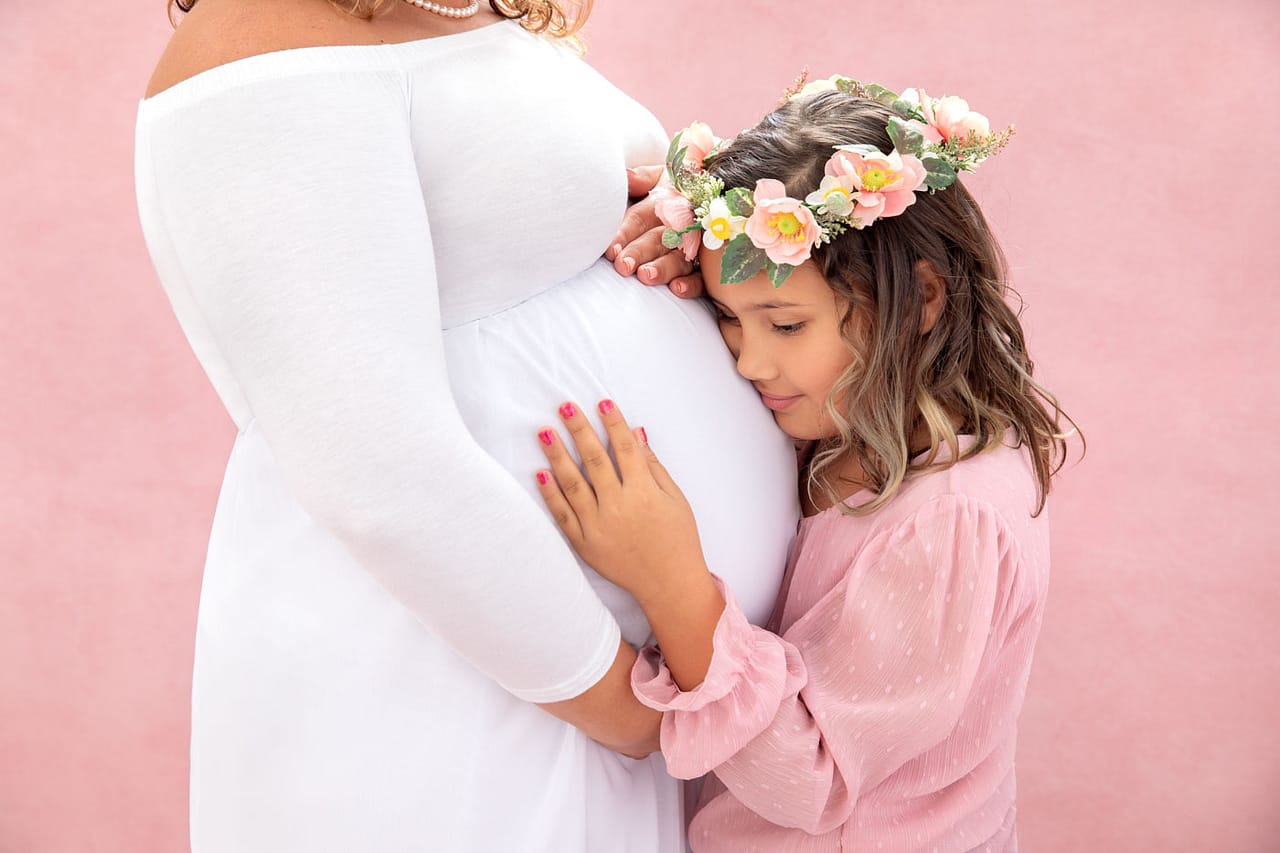 Precious maternity photo of pregnant mother with her daughter. Mother is holding her baby bump and daughter is snuggled close, holding the baby bump. Mother is wearing a white dress and daughter is wearing a pink shirt with a pretty flower crown. Jessica Stellato-Pagan Maternity and Family Portrait Photography.