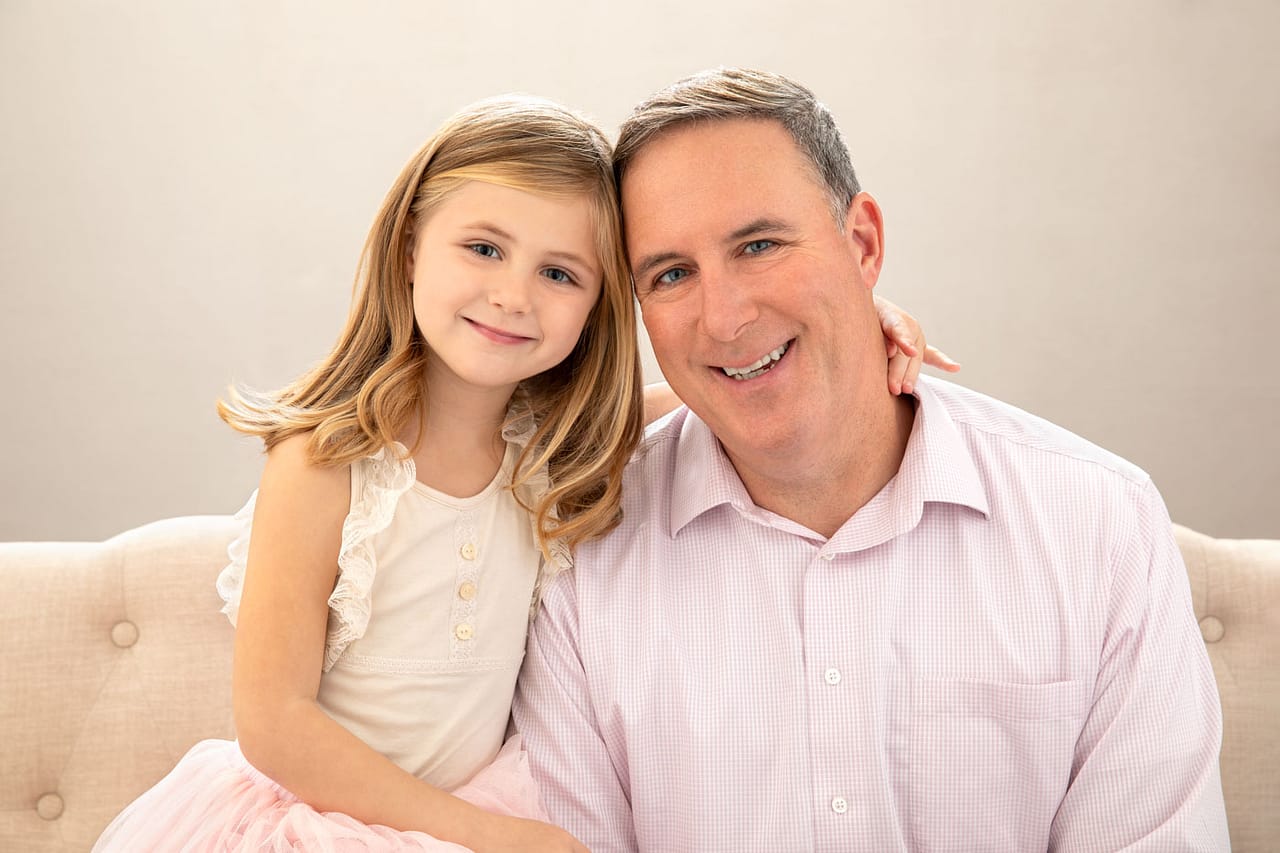 Precious photo of a father and daughter sitting on a couch together. The daughter, who has blonde hair, has her arm around her father's neck. Both are smiling. Chris and Amber Edwards Family Portrait Photography.