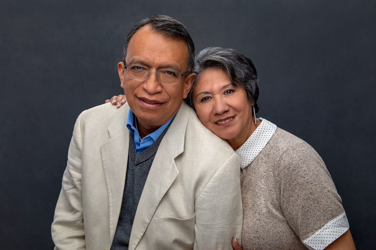 Cute photo of an adorable older couple smiling and standing close, her hand on his shoulder. He's waring a blue shirt, a gray vest and a white jacket. She's wearing a creme heather shirt. Josh and Stefanie Fraley Family Portrait Photography.
