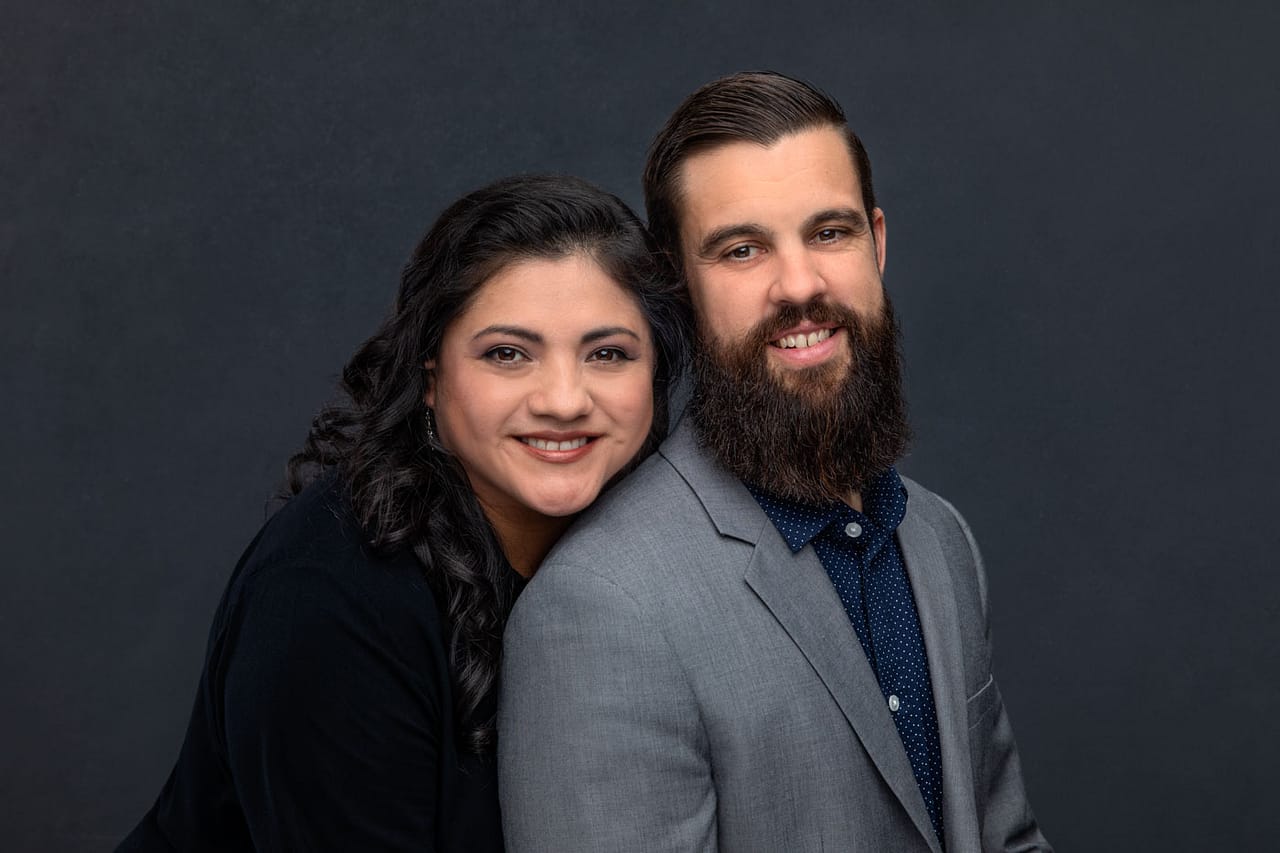 Precious photo of a cute, smiling couple standing close, her head is rested on his shoulder as she stands behind him. He is wearing a blue shirt and gray jacket. She is wearing a black dress. Her hand has a ring and is pulling on his jacket. Josh and Stefanie Fraley Family Portrait Photography.