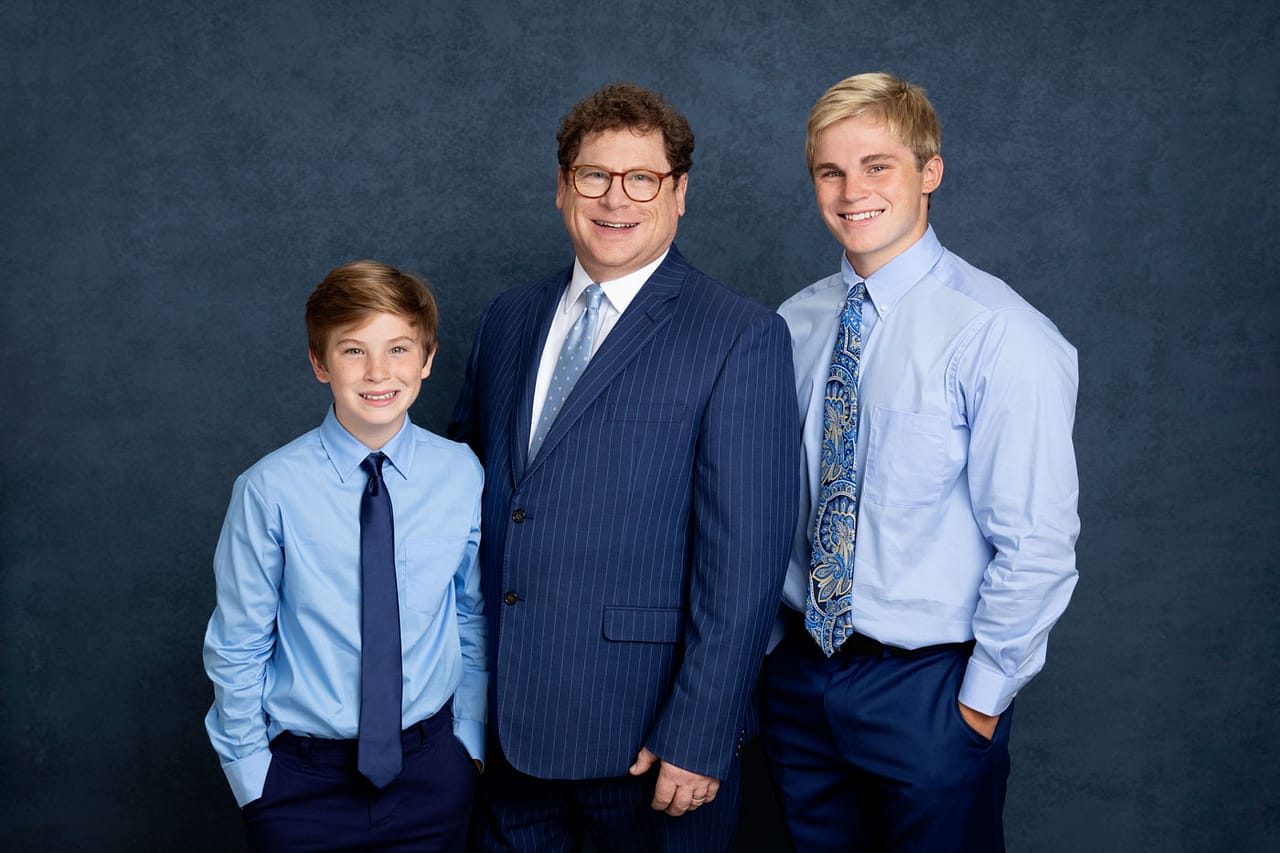 Cute photo of a father and two sons standing and smiling. Father is wearing a blue pin stripe suit and sons are wearing blue shirts with ties. Guy Furay and Myra Ruiz Family Portrait Photography.