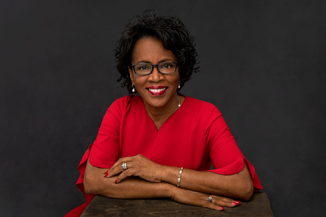 Classy photo of a pretty, smiling black woman with glasses wearing a bright red shirt, black necklace and golden earrings. She's wearing red lipstick and red nails. Janice Butler Women's Portrait Photography.