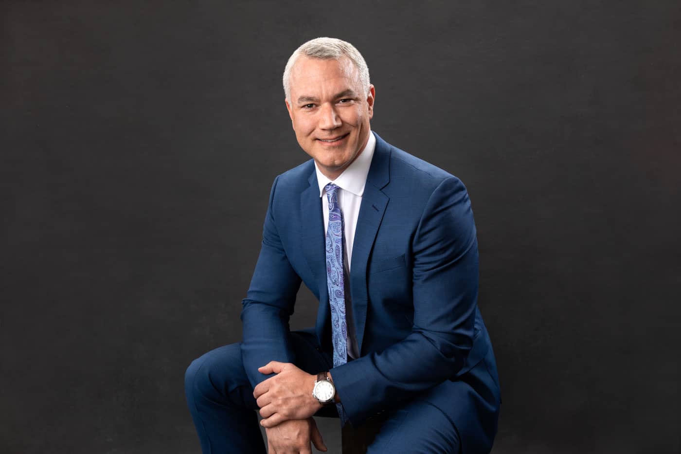 Branding photo of a handsome, stylish man sitting. He's wearing a blue suit and tie and nice watch on a dark gray backdrop. Ben Lovelace Personal Branding Photography