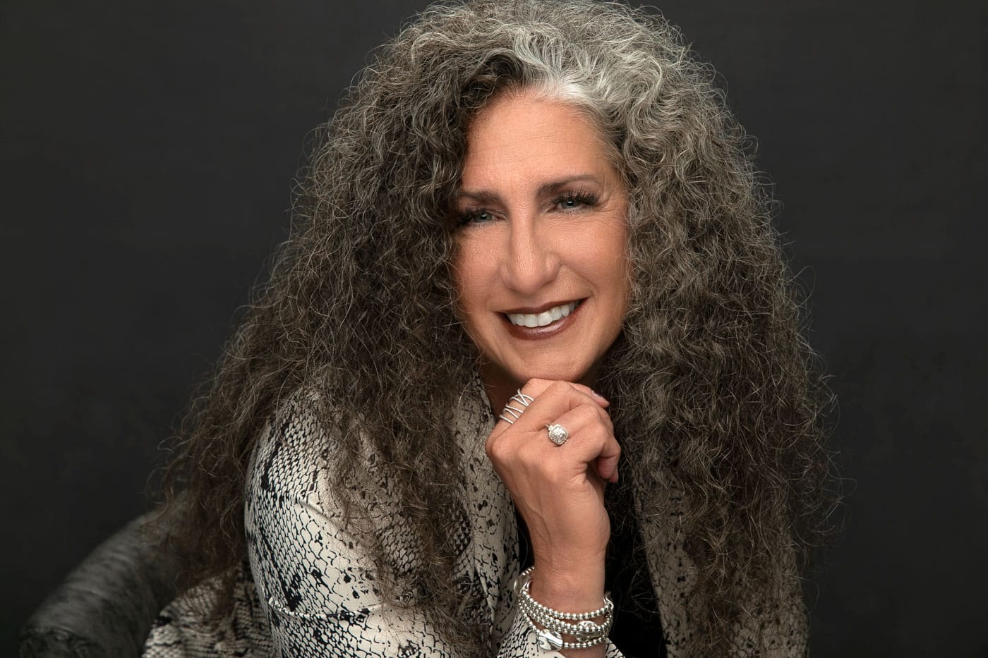 Classy photo of a pretty, smiling woman with pretty wavy salt and pepper hair. She's wearing an aligator print jacket on dark backdrop. Cinnamon McCauley Women's Portrait Photography.