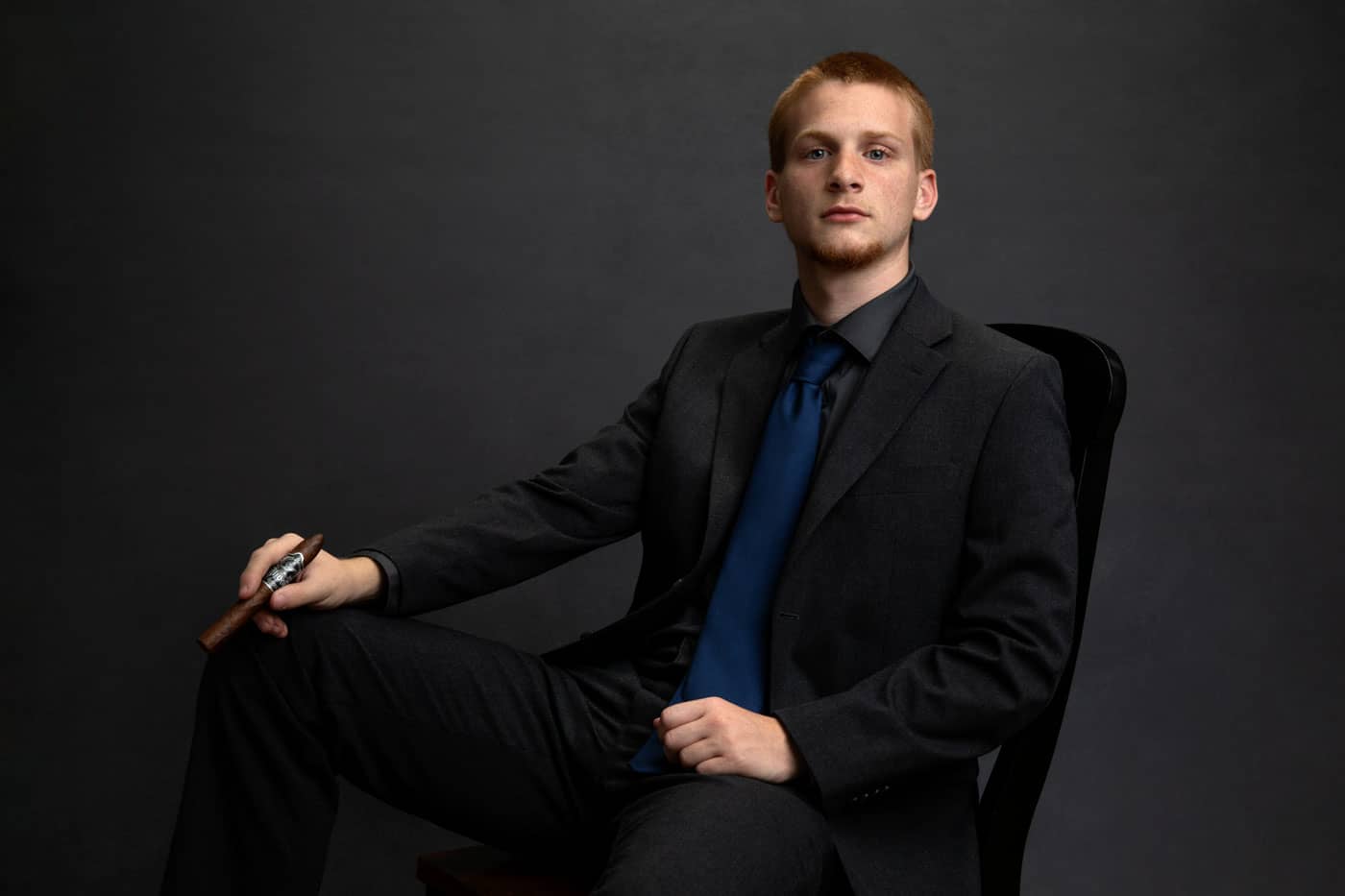 Senior photo of a young man graduating from Greenville High School. He is wearing a black shirt, black jacket and pants with a blue tie. He is sitting in a chair and holding a cigar, a family coming-of-age tradition. Tristan Kopplin Senior Portrait Photography.