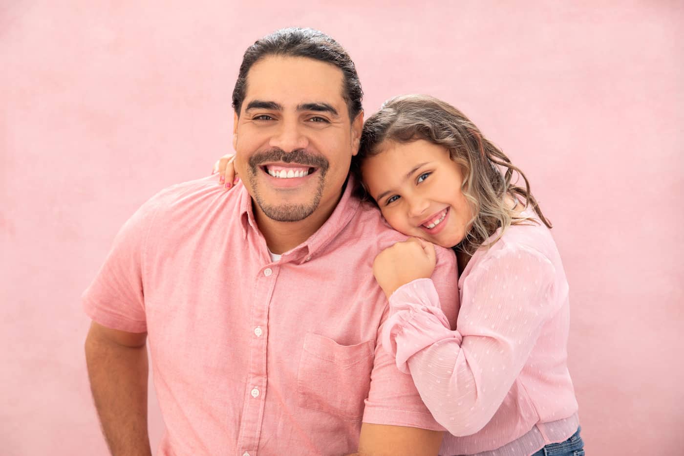 Precious photo of father and daughter. He is sitting and she is snuggled close to him. Both are wearing pink shirts on a pink backdrop. Jessica Stellato-Pagan Maternity and Family Portrait Photography.