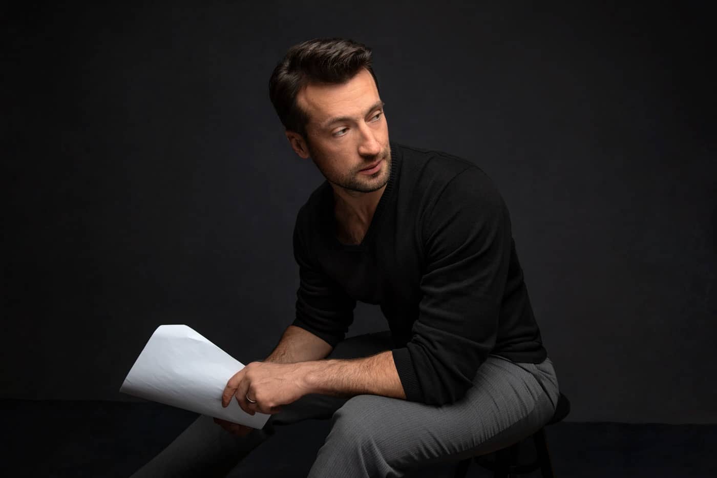 Photo of handsome actor with black shirt and gray pants. He's sitting and working on his lines holding the script. Personal branding photography.