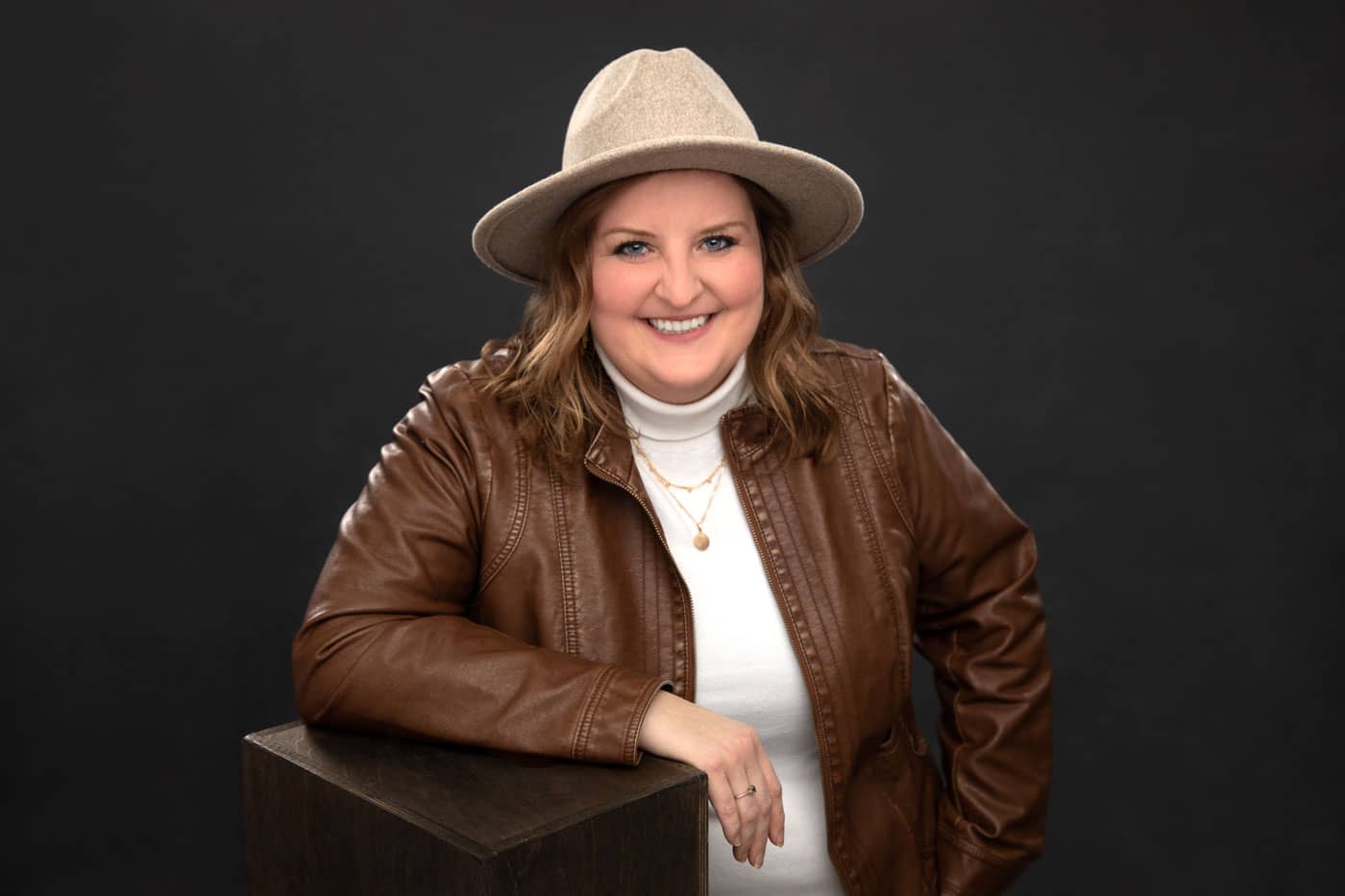Branding photo of a pretty, smiling young woman with brown hair leaning on a box waring a white turtle neck, brown coat and a tan hat. Katie Klaiber Personal Branding Photography.