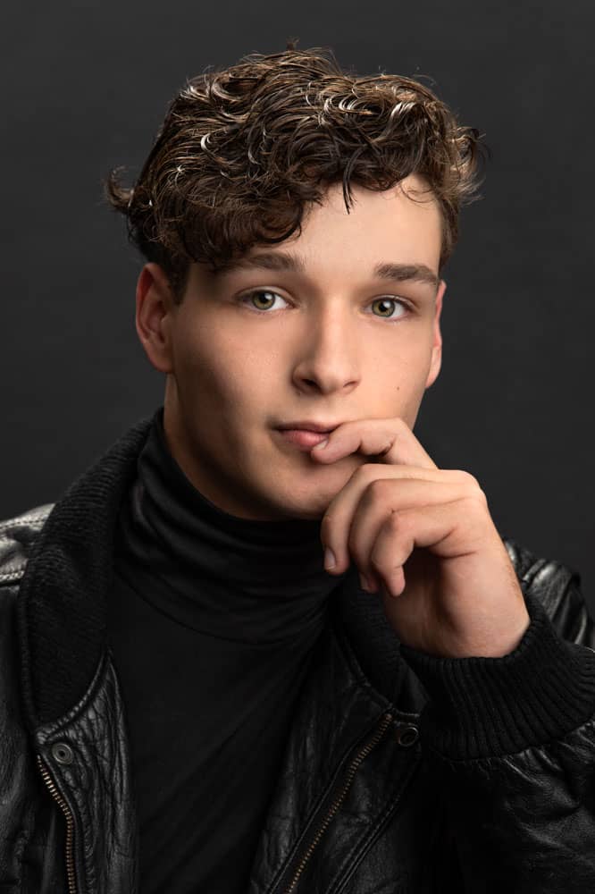Sharp photo of a handsome young man with curly brown hair wearing a black leather jacket. His hand is up with his finger covering his mouth. Lucas Nichter Senior Portrait Photography.