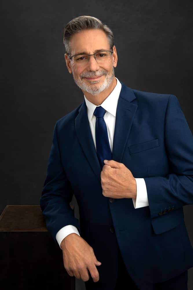 Sharp photo of a handsome man in a blue suit, white shirt and blue tie with a salt and pepper beard and glasses. Donald Aucoin engagemnt portrait photography.