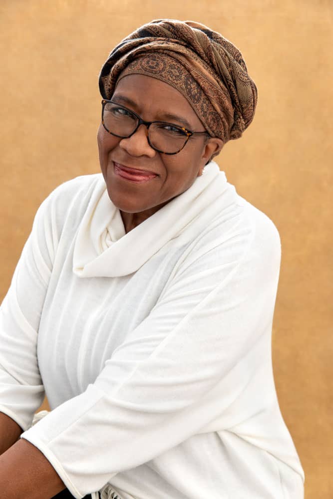 Classy photo of pretty black woman smiling. She's wearing a brown, textured head covering and a white shirt on tan backdrop. Audra Odom Women's Portrait Photography.