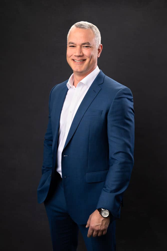 Branding photo of a handsome, stylish man standing. He's wearing a blue suit and tie and nice watch on a dark gray backdrop. Ben Lovelace Personal Branding Photography