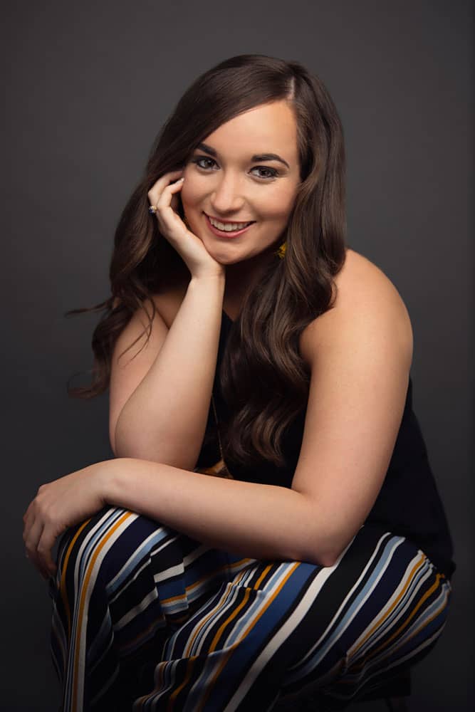 Senior photo of a pretty, young smiling woman sitting on a stool, leaning forward, chin in her hand. She's wearing striped pants on a dark backdrop. Chelsea Bergeron Senior Portrait Photography.