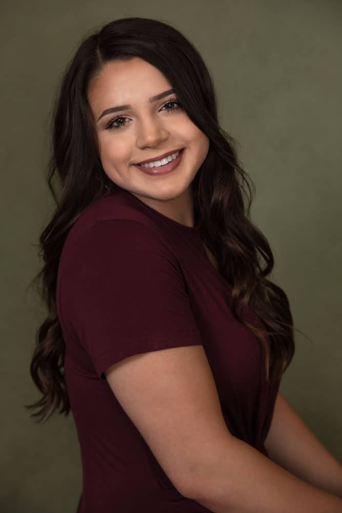 Senior photo of a pretty, smiling young woman with a maroon shirt and lipstick on olive backdrop. High school senior. Denee Dhuet Senior Portrait Photography.