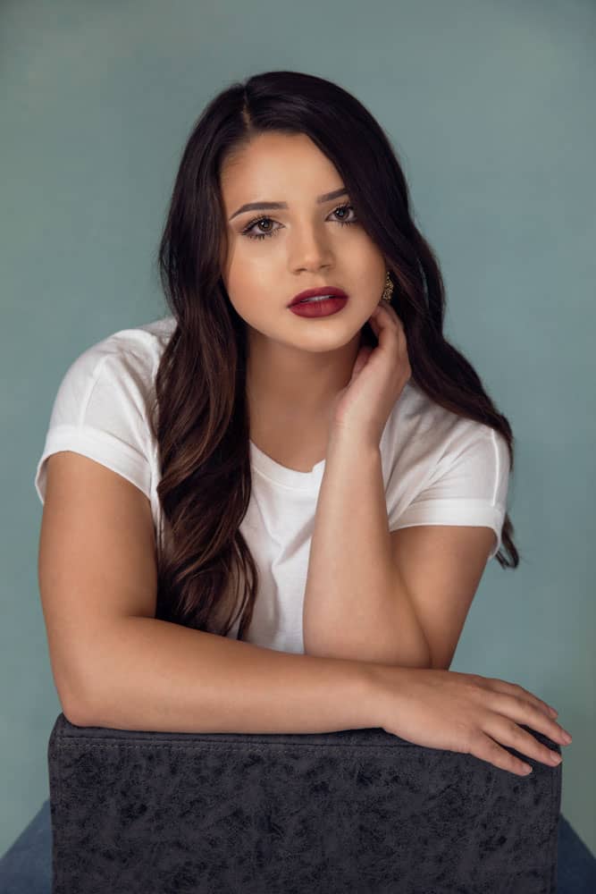 Senior photo of a pretty, smiling young woman with white shirt and red lipstick on blue backdrop. High school senior. Denee Dhuet Senior Portrait Photography.