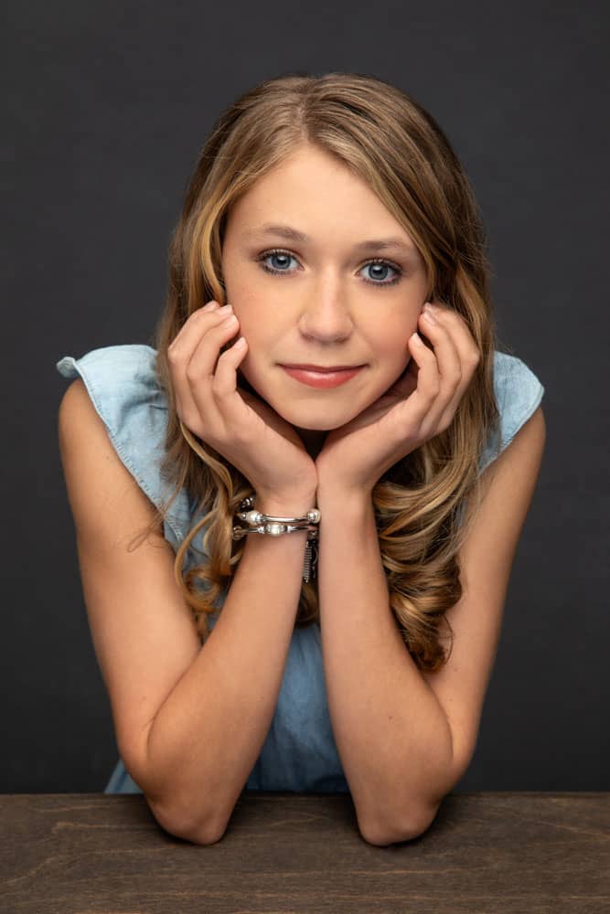 Cute photo of a pretty, young woman leaning forward with her chin in her hands, wearing a blue dress. Don and Beverly Elmore Family Portrait Photography.