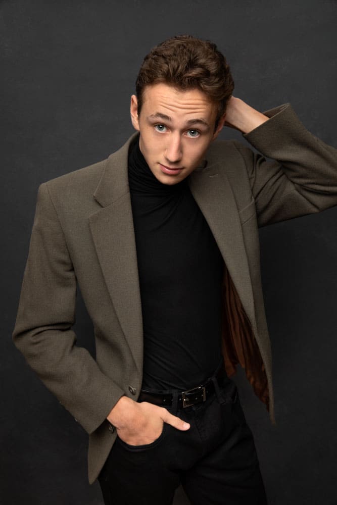 Slick photo of a handsome high school senior with a muted green jacket and black shirt. One hand is in his pocket the other touching the back of his head. Elmore Senior Portrait Photography.