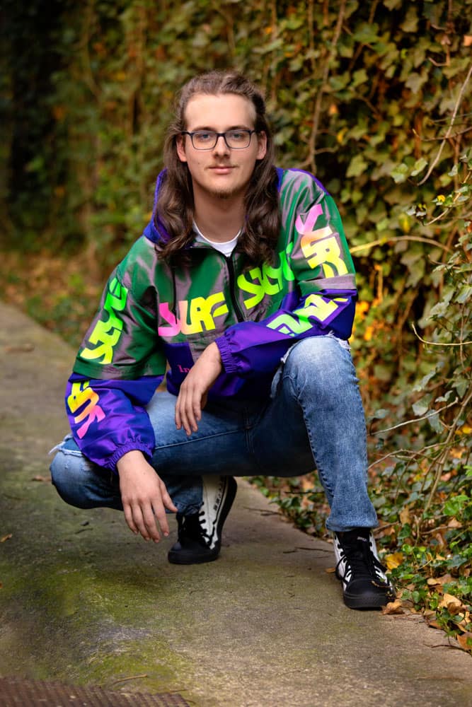 Senior photo of a smiling young man with long brown hair and glasses. He's wearing an iconic Surf Style bomber jacket and crouching down in front of an ivy wall. Max Anderson Senior Portrait Photography.