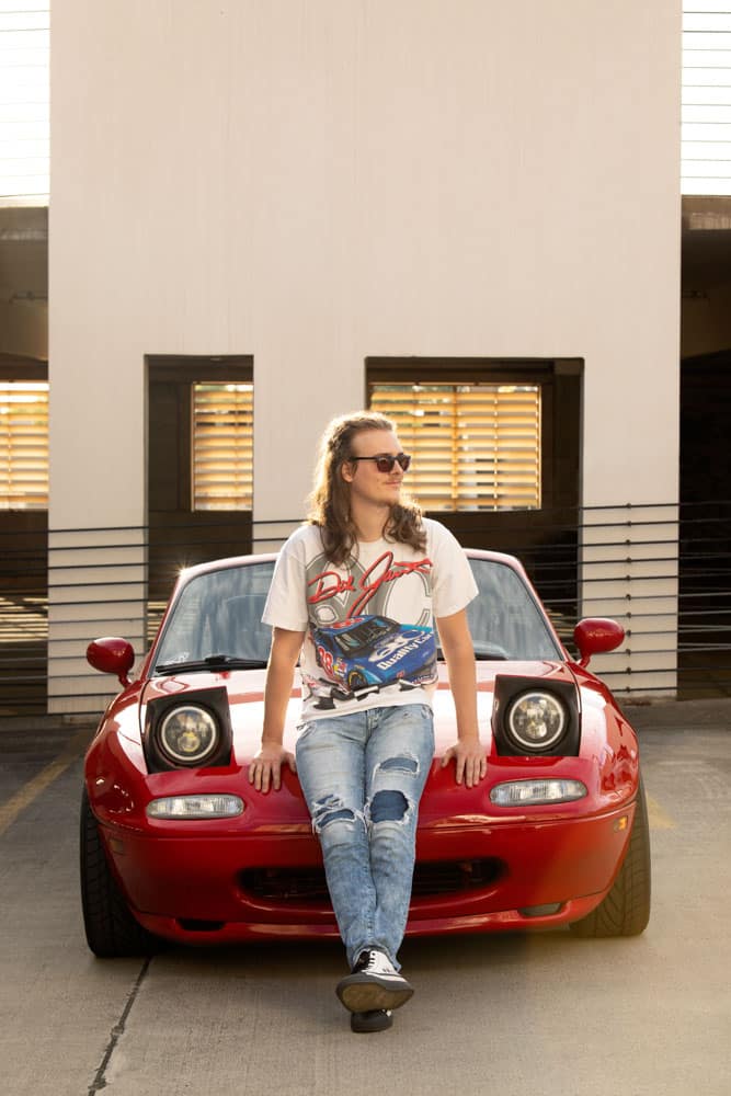 Senior photo of a young man with long brown hair and glasses. He's wearing white graphic tee as he leans on the hood of his red 1990 Mazda Miata. Max Anderson Senior Portrait Photography.
