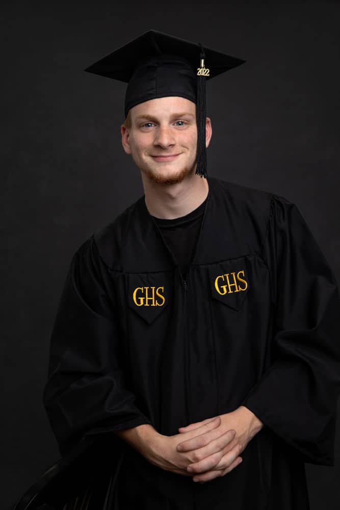 Senior photo of a young man graduating from Greenville High School. He is wearing a black cap and gown, with bright GHS letters embroidered on his gown. Tristan Kopplin Senior Portrait Photography.