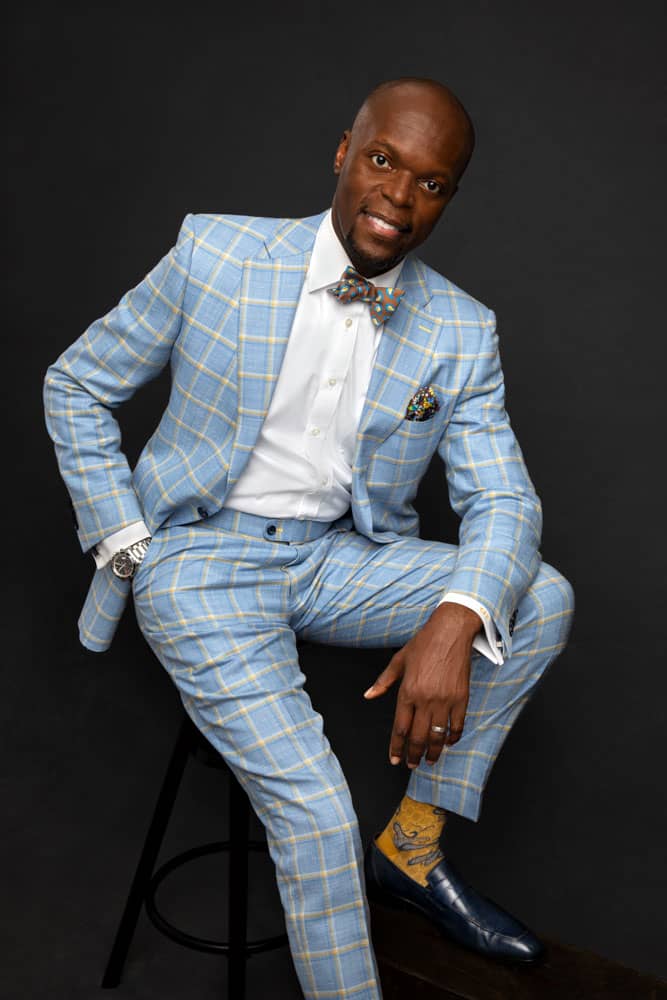 Branding photo of a stylish, handsome black man sitting on a stool, elbow resting on his knee. He's wearing a sky blue suit with paisley bowtie, floral pocket square, yellow socks and dark blue leather shoes. Charles Davis of A Polished Man during personal branding photo session.