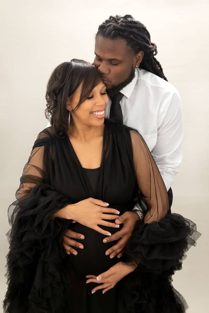 Precious maternity photo of a cute couple. They are standing close; with the husband's hands around his wife's belly; and her hands on her belly. She shows her pretty smile and looks down as he kisses her forehead. She is wearing a long black tulle maternity gown and he is wearing a white shirt with a black tie. Holmes Family Portrait Photography.